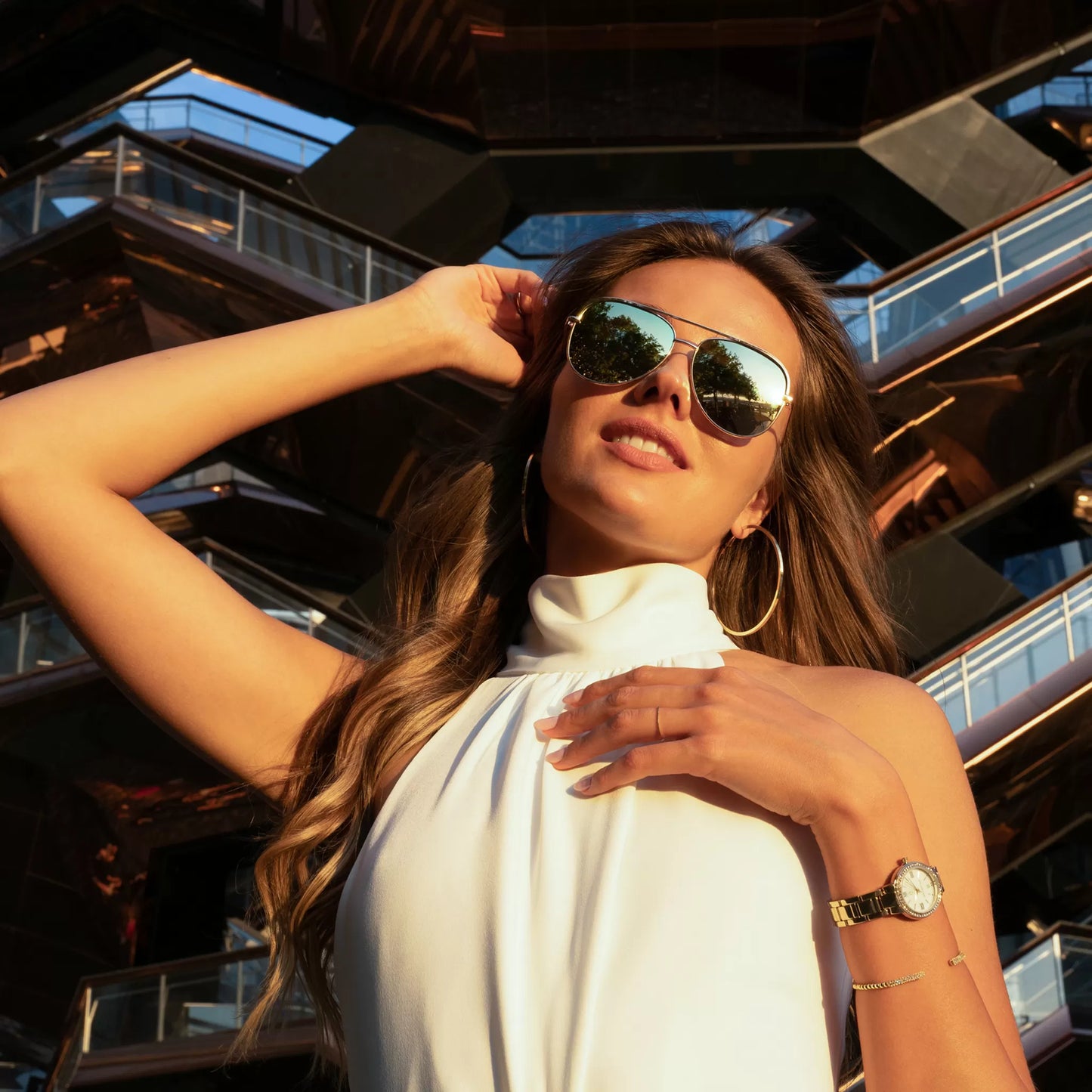 Model wearing Monarchic® Watch - Cosmopolitan - Jackie Edition - Gold link watch and gold accessories, large gold hoop earrings and aviator glasses posing in front of Hudson Yard's Vessel